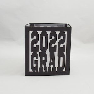 May include: A black square box with the white text "2022 GRAD" cut out on the front. The box is likely made of cardstock or a similar material. The background is plain white.