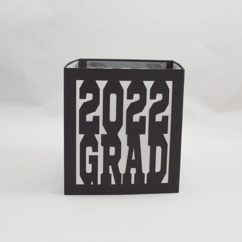 May include: A black square box with the white text "2022 GRAD" cut out on the front. The box is likely made of cardstock or a similar material. The background is plain white.