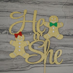 May include: Gold glitter cake topper with the words "He or She" and two gingerbread people. One gingerbread person has a red bow, and the other has a green bow tie. The topper is on a wooden stick.