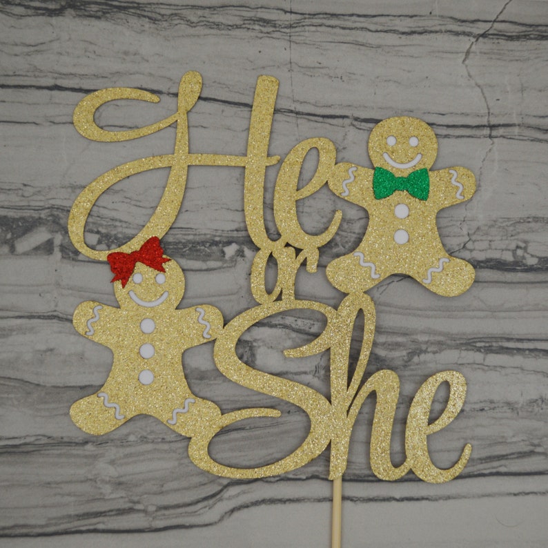 May include: Gold glitter cake topper with the words "He or She" and two gingerbread people. One gingerbread person has a red bow, and the other has a green bow tie. The topper is on a wooden stick.