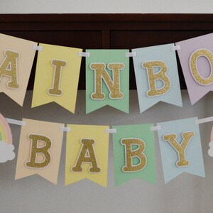 May include: A pastel rainbow baby banner with gold glitter letters. The banner says "Rainbow Baby" and has two rainbow cutouts with clouds on either end.