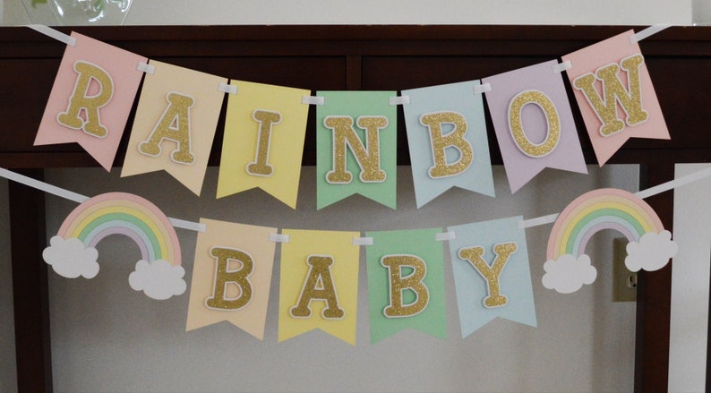 May include: A pastel rainbow baby banner with gold glitter letters. The banner says "Rainbow Baby" and has two rainbow cutouts with clouds on either end.