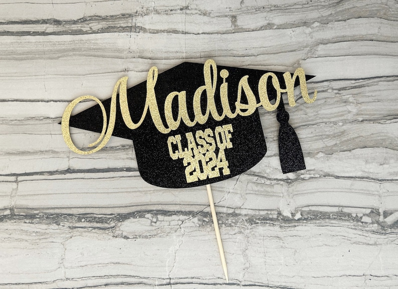 Graduation cap shape glitter cake topper personalized with name across the top and Class of 2024 along the bottom. Shown here with the name Madison in gold glitter on a black graduation cap.