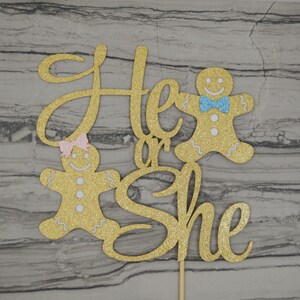 May include: A gold glitter cake topper with the words "He or She" in a cursive font. Two gingerbread people are on either side of the text, one with a pink bow and the other with a blue bow. The topper is attached to a wooden stick.