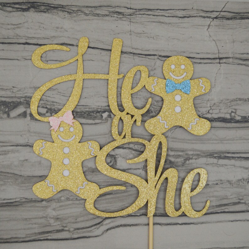 May include: A gold glitter cake topper with the words "He or She" in a cursive font. Two gingerbread people are on either side of the text, one with a pink bow and the other with a blue bow. The topper is attached to a wooden stick.