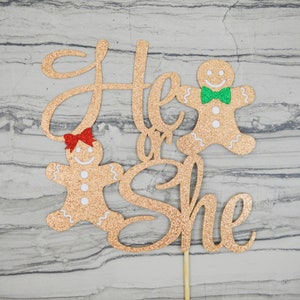 May include: A glittery gold cake topper with the words "He or She" in cursive script. Two gingerbread people are on either side of the text, one with a red bow and the other with a green bow tie. The topper is attached to a wooden stick.