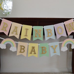 May include: A pastel rainbow baby banner with gold glitter letters. The banner has two rainbow and cloud cutouts on either end.