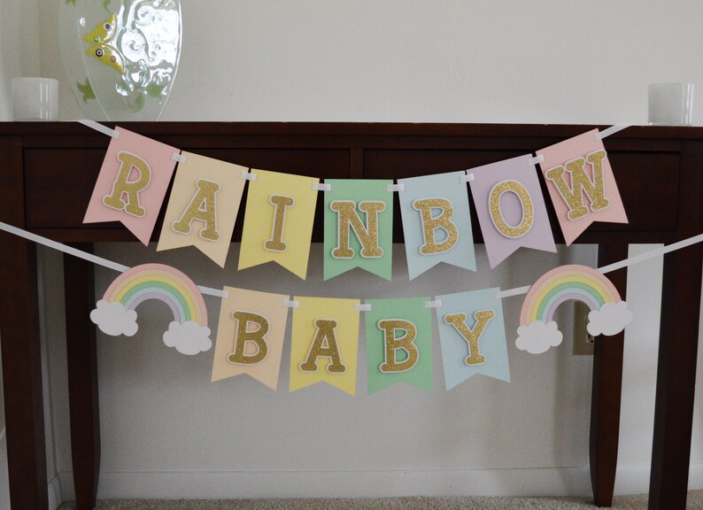 May include: A pastel rainbow baby banner with gold glitter letters. The banner has two rainbow and cloud cutouts on either end.