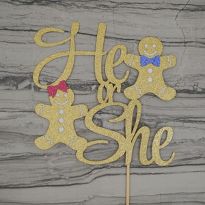 May include: A gold glitter cake topper with the words "He or She" in cursive script. Two gingerbread people are on either side of the text. One has a pink bow, and the other has a blue bow. The topper is attached to a wooden stick.