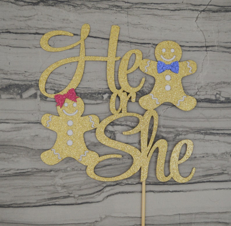 May include: A gold glitter cake topper with the words "He or She" in cursive script. Two gingerbread people are on either side of the text. One has a pink bow, and the other has a blue bow. The topper is attached to a wooden stick.