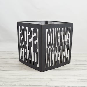 May include: A black, square graduation box with cut-out text. The text reads "2022 GRAD" on one side and "CONGRATS MCKENZIE" on another. The box is made of a dark material and has a white interior, creating a contrast with the cut-out letters.