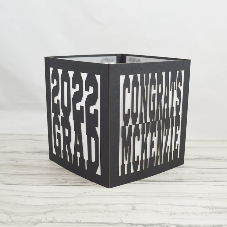 May include: A black, square graduation box with cut-out text. The text reads "2022 GRAD" on one side and "CONGRATS MCKENZIE" on another. The box is made of a dark material and has a white interior, creating a contrast with the cut-out letters.
