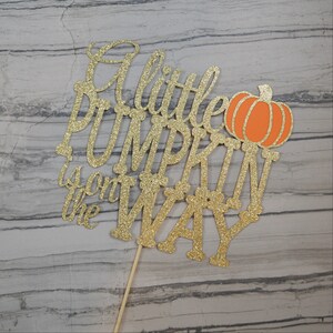 Puede incluir: Adorno para tarta dorado con purpurina con la frase "A little PUMPKIN is on the WAY" y una ilustraci&oacute;n de calabaza naranja. El adorno est&aacute; sujeto a un palito de madera, ideal para un baby shower.