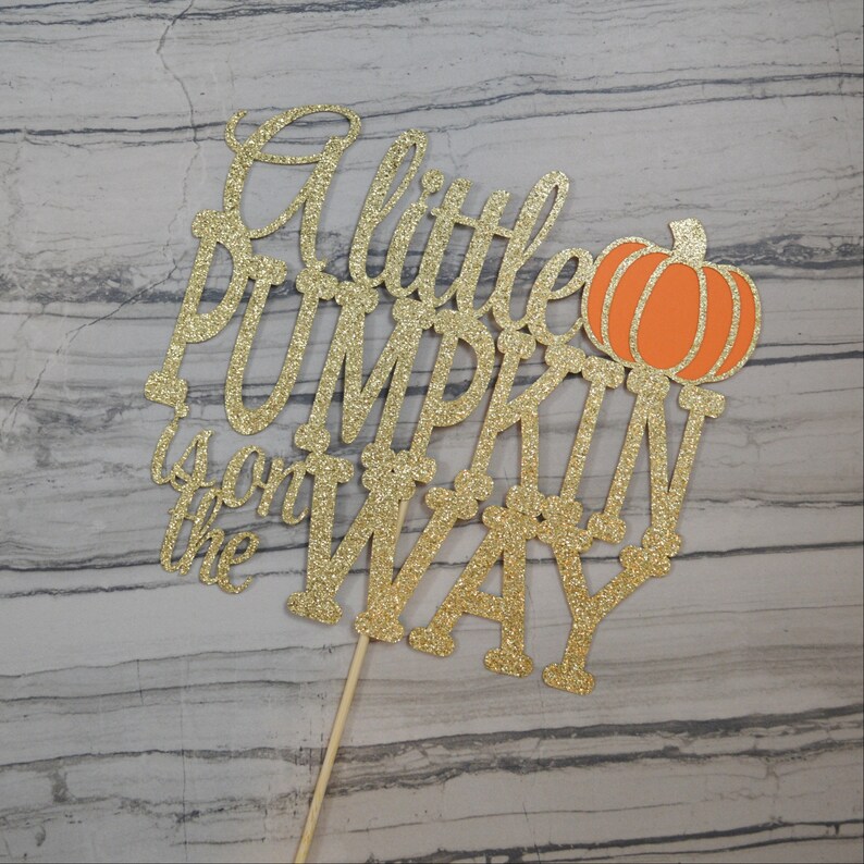 Puede incluir: Adorno para tarta dorado con purpurina con la frase "A little PUMPKIN is on the WAY" y una ilustraci&oacute;n de calabaza naranja. El adorno est&aacute; sujeto a un palito de madera, ideal para un baby shower.