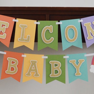May include: A rainbow-colored banner with the words "WELCOME BABY" in gold glitter letters. The banner is strung with white ribbon and has rainbow-colored clouds on either end.