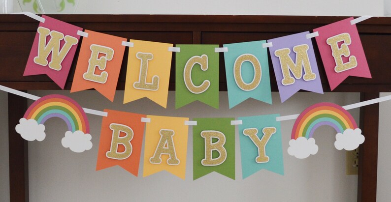 May include: A rainbow-colored banner with the words "WELCOME BABY" in gold glitter letters. The banner is strung with white ribbon and has rainbow-colored clouds on either end.