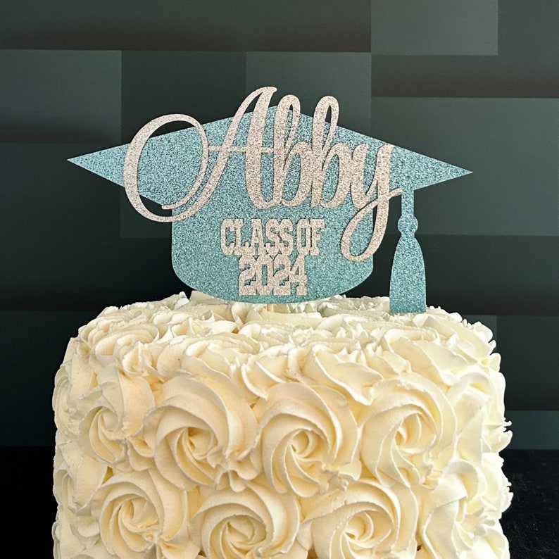 Graduation cap shape glitter cake topper personalized with name across the top and Class of 2024 along the bottom. Shown here with the name Abby in silver glitter on a light blue graduation cap displayed on a white cake.