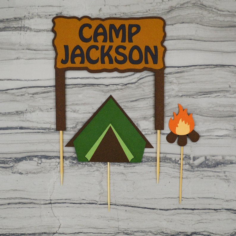 May include: A set of three cake toppers featuring a brown sign with the text "Camp Jackson", a green tent, and a campfire with orange flames. The toppers are made of felt and are attached to wooden skewers.