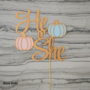 May include: Rose gold glitter cake topper with the words "He or She" and two pumpkins, one pink and one blue. The topper is attached to a wooden stick for easy insertion into a cake.