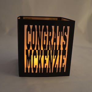 May include: A black paper lantern with the words "CONGRATS MCKENZIE" cut out. The lantern is lit from within, creating a warm, inviting glow. The design is simple yet elegant, perfect for a celebration.