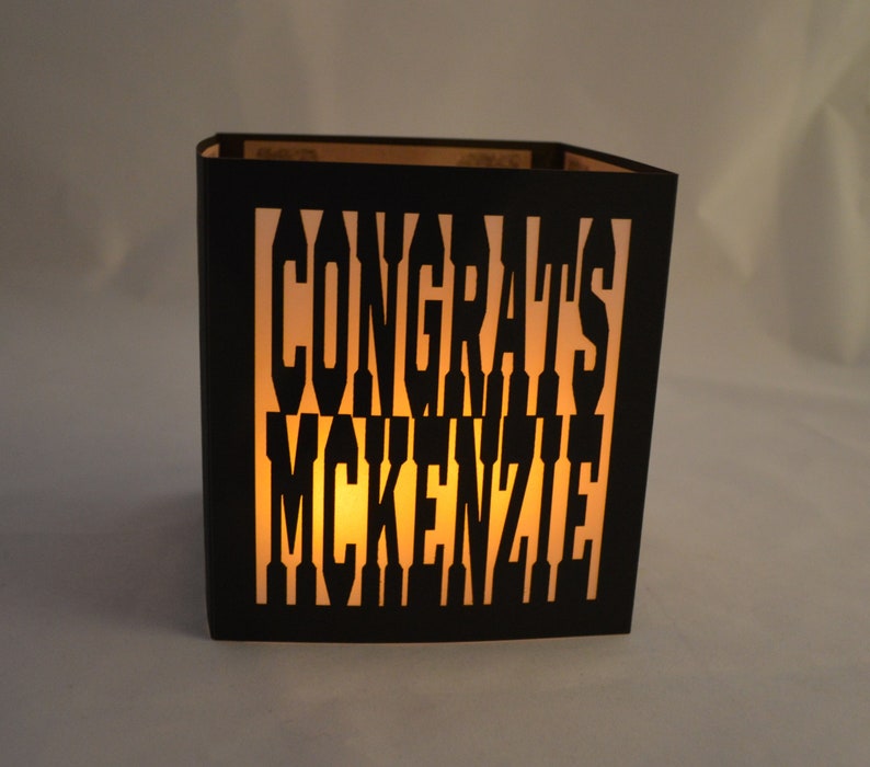May include: A black paper lantern with the words "CONGRATS MCKENZIE" cut out. The lantern is lit from within, creating a warm, inviting glow. The design is simple yet elegant, perfect for a celebration.