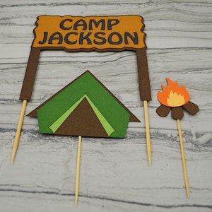 May include: A set of three cake toppers featuring a brown sign with the text "Camp Jackson", a green tent, and a brown campfire with orange flames.