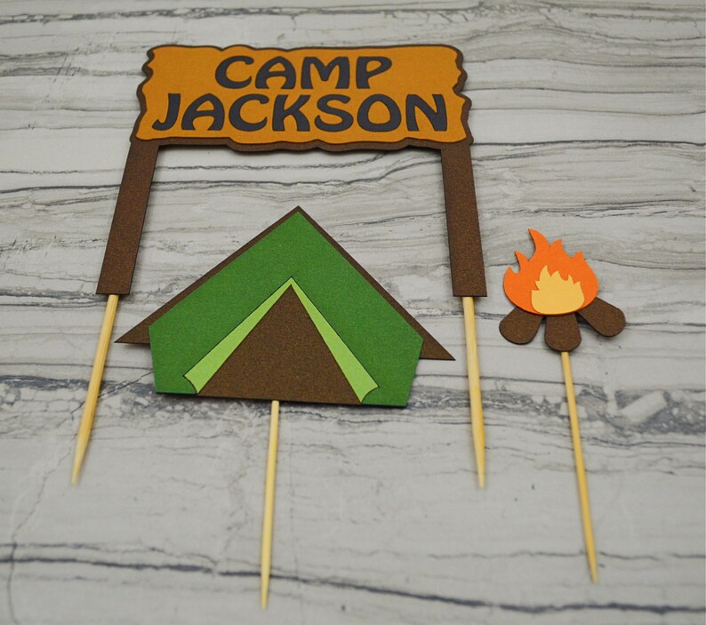 May include: A set of three cake toppers featuring a brown sign with the text "Camp Jackson", a green tent, and a brown campfire with orange flames.