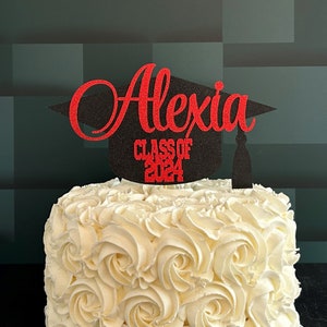 Graduation cap shape glitter cake topper personalized with name across the top and Class of 2024 along the bottom. Shown here with the name Alexia in red glitter on a black graduation cap displayed on a white cake.