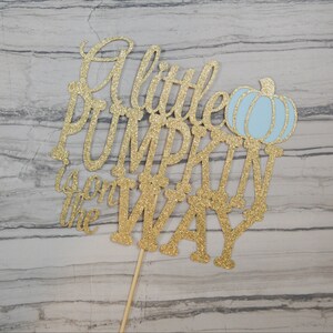 Puede incluir: Un adorno para tarta de purpurina dorada con la frase "A little PUMPKIN is on the WAY" y una calabaza azul claro. El adorno est&aacute; sujeto a un palito de madera.
