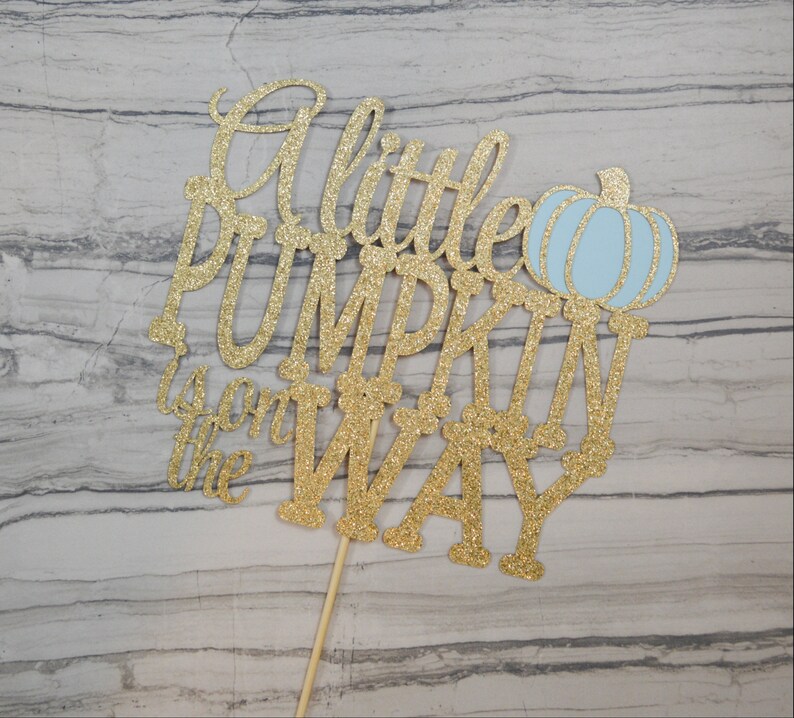 Puede incluir: Un adorno para tarta de purpurina dorada con la frase "A little PUMPKIN is on the WAY" y una calabaza azul claro. El adorno est&aacute; sujeto a un palito de madera.