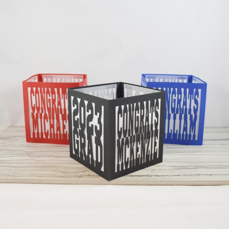 May include: Three colourful graduation-themed paper lanterns. One is red with "CONGRAT MICHAEL" cut out, another is black with "2023 GRAD CONGRATS MCKENZIE", and the third is blue with "CONGRATS WILLIAM".