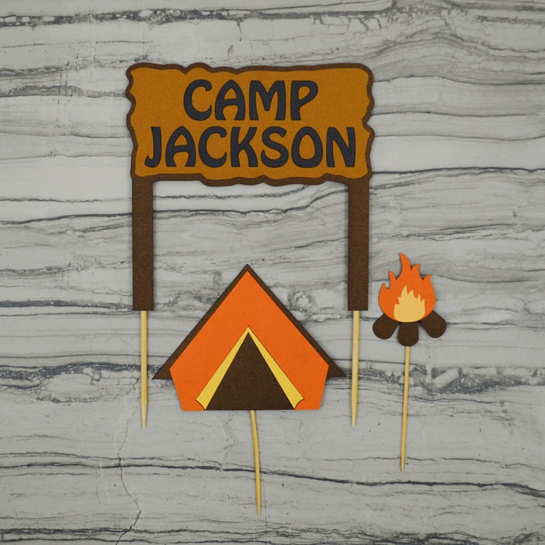 May include: A set of three cake toppers featuring a brown sign with the text "Camp Jackson", a brown and orange tent, and a campfire with orange flames. The toppers are on wooden skewers.