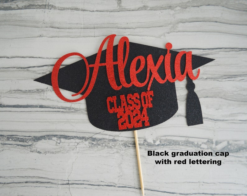 Graduation cap shape glitter cake topper personalized with name across the top and Class of 2024 along the bottom. Shown here with the name Alexia in red glitter on a black graduation cap.