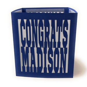 May include: A blue paper card with the words "CONGRATS MADISON" cut out. The card is a rectangular shape with a white interior, and the text is visible through the cut-out design.