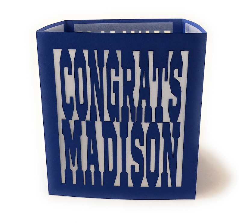 May include: A blue paper card with the words "CONGRATS MADISON" cut out. The card is a rectangular shape with a white interior, and the text is visible through the cut-out design.
