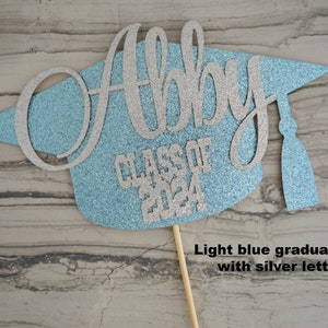 Graduation cap shape glitter cake topper personalized with name across the top and Class of 2024 along the bottom. Shown here with the name Abby in silver glitter on a light blue graduation cap.