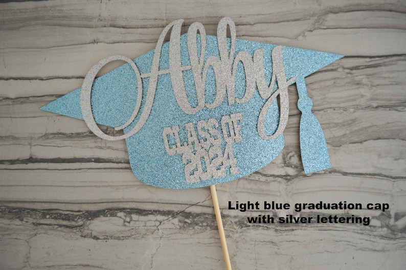 Graduation cap shape glitter cake topper personalized with name across the top and Class of 2024 along the bottom. Shown here with the name Abby in silver glitter on a light blue graduation cap.