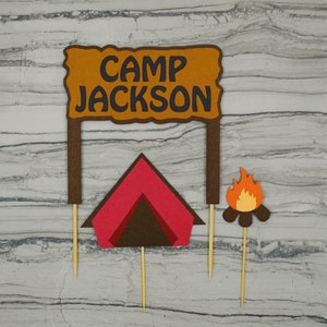 May include: Three brown cardboard cake toppers with a red and brown tent, a campfire with orange flames, and a sign that reads "Camp Jackson".