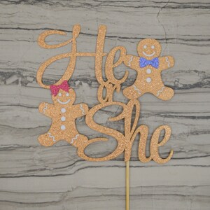 May include: A glittery gold cake topper with the words "He or She" in cursive script. Two gingerbread people are featured, one with a pink bow and the other with a blue bow tie. The topper is attached to a wooden stick.