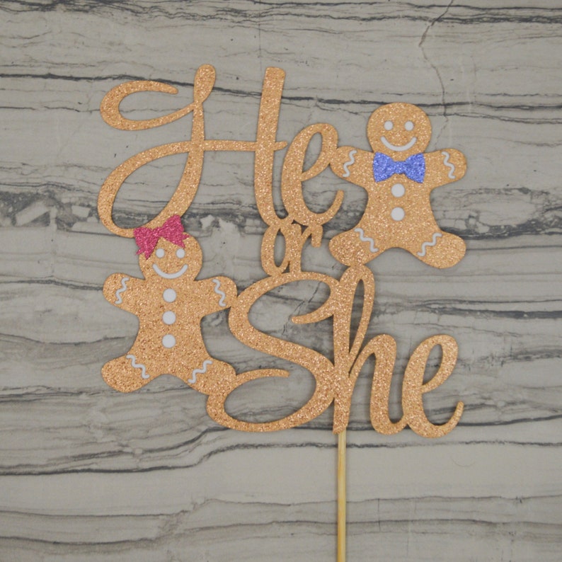 May include: A glittery gold cake topper with the words "He or She" in cursive script. Two gingerbread people are featured, one with a pink bow and the other with a blue bow tie. The topper is attached to a wooden stick.