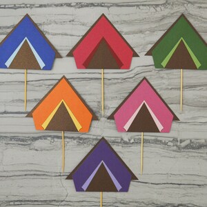 May include: Six colorful paper tent cupcake toppers. The tents are made of different colors, including blue, red, green, orange, pink, and purple. Each tent has a brown flap and a white or yellow interior.