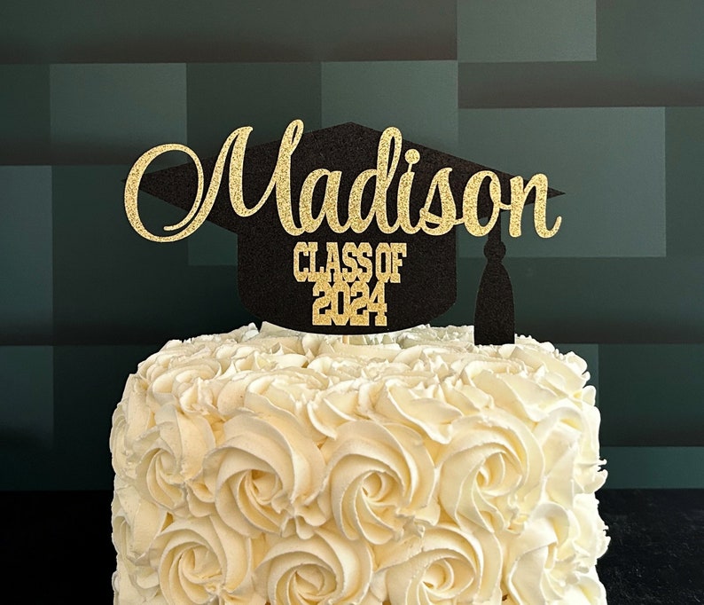 Graduation cap shape glitter cake topper personalized with name across the top and Class of 2024 along the bottom. Shown here with the name Madison in gold glitter on a black graduation cap displayed on a white cake.