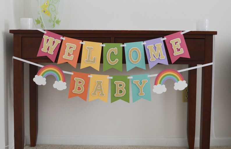 May include: A rainbow-themed baby shower banner with the words "Welcome Baby" in gold glitter letters. The banner is strung on a white ribbon and has two rainbow and cloud decorations.