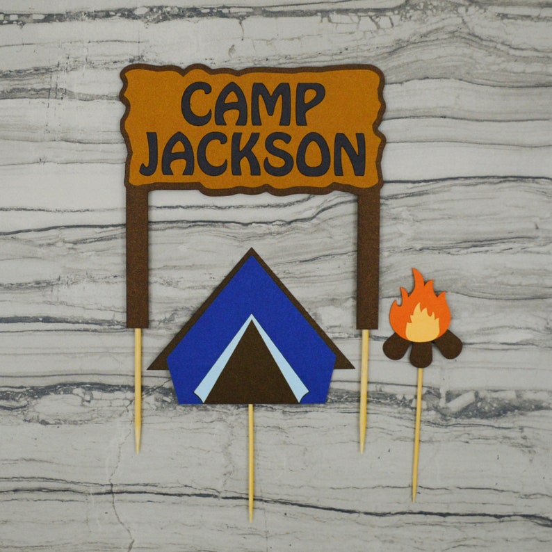 May include: A set of three cake toppers featuring a brown sign with the text "Camp Jackson", a blue tent, and a campfire with orange flames. The toppers are made of felt and are attached to wooden skewers.