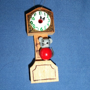 May include: Wooden Christmas ornament shaped like a grandfather clock. The clock face is white with green and black accents. A gray mouse with a red ball body is in front of the clock. The ornament hangs from a gold string.