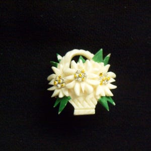 May include: A cream-colored floral brooch featuring a basket of flowers. The flowers have white petals and yellow centers, with green leaves. The brooch is set against a black background.