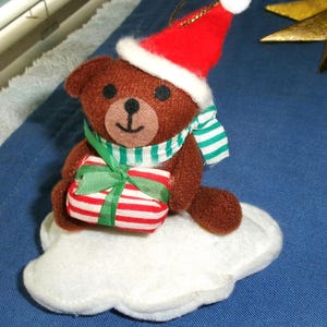 May include: A brown teddy bear Christmas ornament wearing a red Santa hat and a green and white striped scarf. The bear holds a wrapped gift with red and white stripes and a green bow, sitting on a white cloud.
