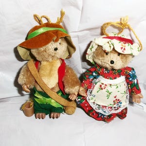 May include: Two plush teddy bears dressed in vintage-style outfits. One bear is wearing a brown hat with a green band and a green vest. The other bear is wearing a red and white floral dress with a white apron.