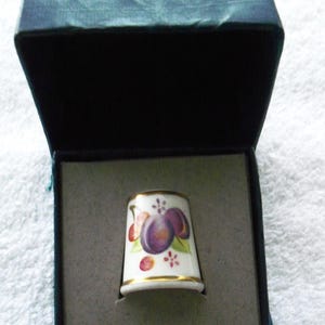 May include: A Royal Worcester Bone China thimble in a presentation box. The thimble is white with a gold rim and features a plum design. The box is dark blue with the words "Royal Worcester Bone China" printed on the lid.