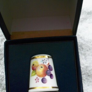 May include: A white porcelain thimble with a gold trim and a colorful fruit design. The thimble is in a blue box.